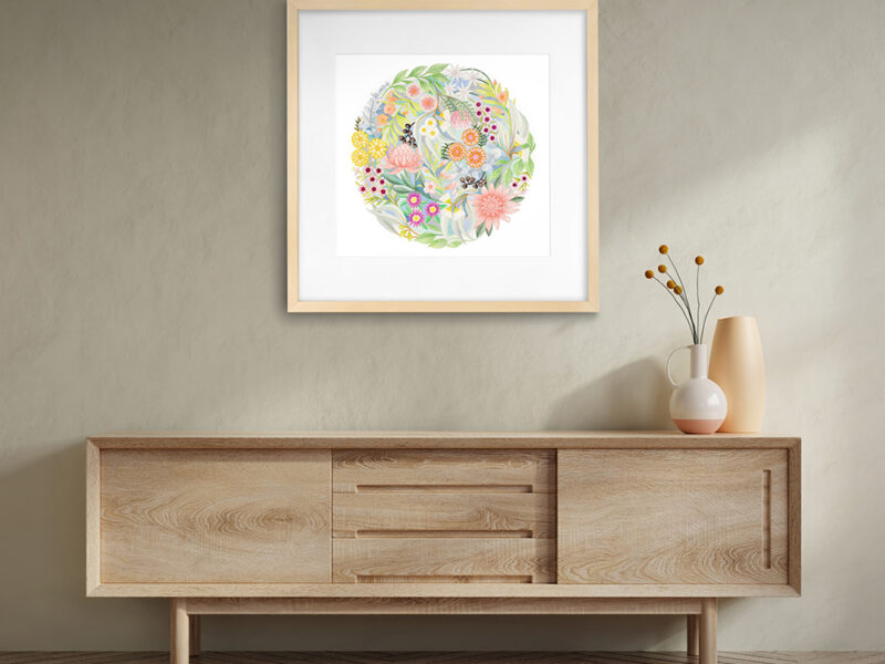 A multi-coloured circular artwork by Claire Ishino in a square frame hangs on a neutral coloured wall with wooden sideboard beneath and vase of flowers on the right.