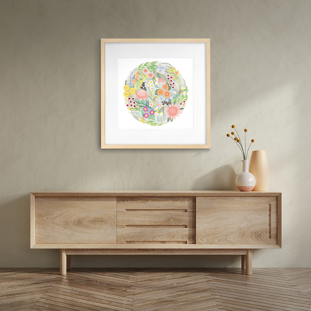 A multi-coloured circular artwork by Claire Ishino in a square frame hangs on a neutral coloured wall with wooden sideboard beneath and vase of flowers on the right.